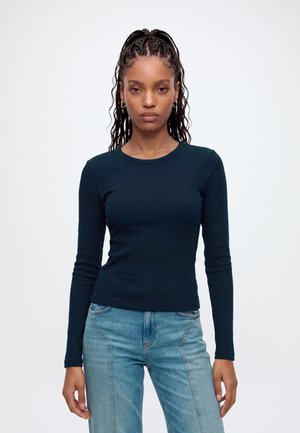 Ung kvinde med flettet hår bærer guldfarvede hoops-øreringe, en tætsiddende mørkeblå langærmet top og lyseblå højttaljede jeans mod en ensfarvet baggrund.