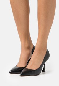 Escarpins noirs en cuir avec un bout pointu, texture lisse et un design de talon carré distinctif. Silhouette minimaliste et épurée.