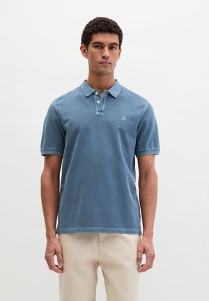 Mann trägt blaues kurzärmeliges Polohemd und beige Hose, steht vor einem schlichten hellen Hintergrund.