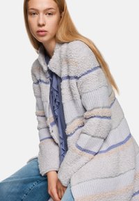 Long, textured cardigan in light gray and cream, featuring blue and beige stripes. Styled with a ruffled blue blouse and denim jeans.