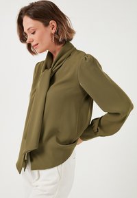 Olive green blouse with a tied collar, puffed sleeves, soft fabric, and a curved hem. Paired with white pants, showcasing a relaxed fit.