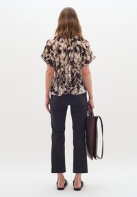 Doorzichtig top met abstract patroon in crème, zwart en bruin, in combinatie met strakke zwarte broeken. Houdt een bruine leren tas vast.