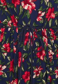 Robe fleurie avec un fond marine orné de grandes fleurs d'hibiscus rouges et de feuilles vertes, cintrée à la taille par une ceinture tressée.