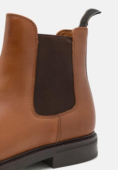 Bota Chelsea de couro castanho com painel lateral elástico, material stretch escuro em contraste e puxador preto; sola plana com detalhes texturizados.