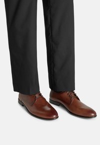 Hnědé kožené společenské boty s kulat�ými špičkami, tenkými šňůrkami a perforovanými detaily, v kombinaci s černými kalhotami. Na kůži se odráží lesk.