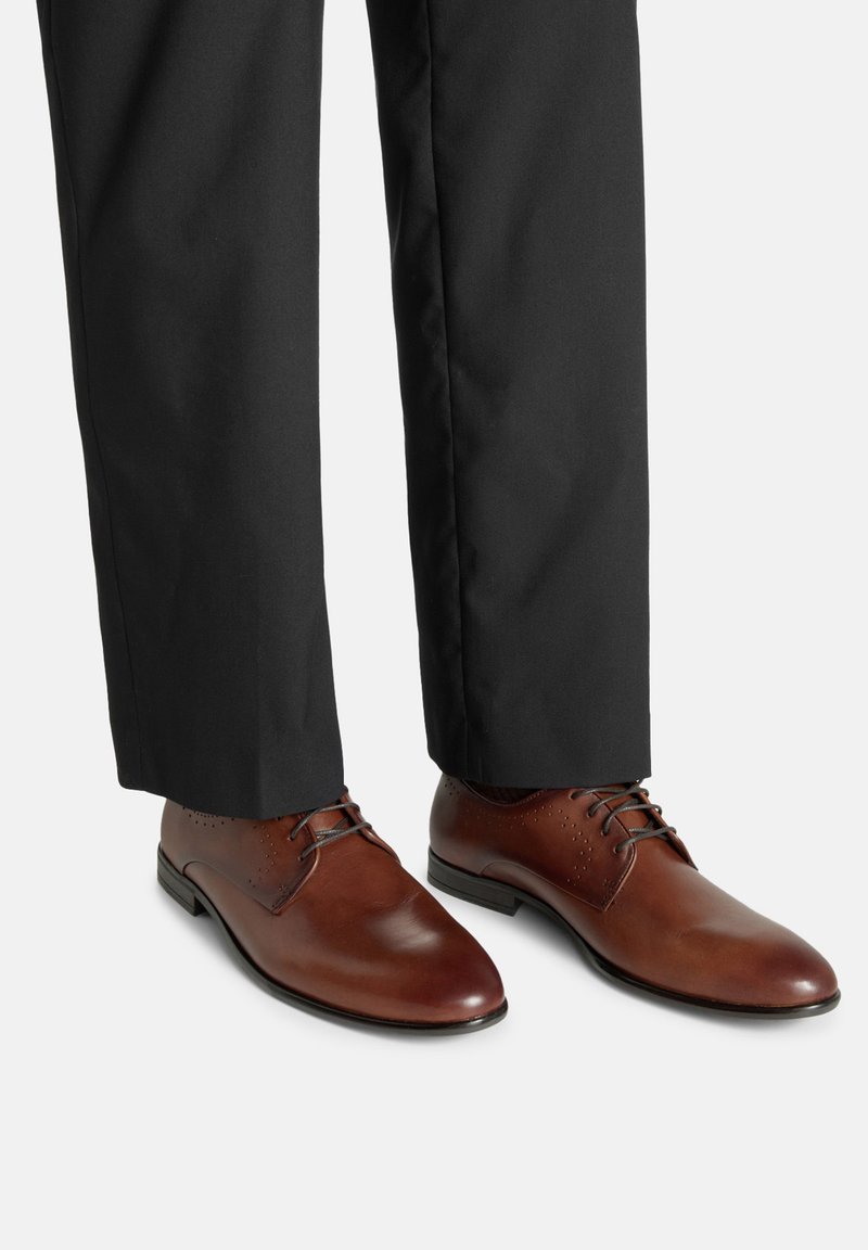 Hnědé kožené společenské boty s kulatými špičkami, tenkými šňůrkami a perforovanými detaily, v kombinaci s černými kalhotami. Na kůži se odráží lesk.