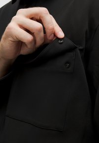 Must musta tekstuuriga riie, millel on rinna-tasku. Käsi avab taskut, millel on rõngasnuppe sulgemiseks.