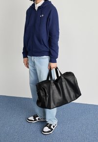 Bolsa de viaje negra de piel con dos asas, textura suave, forma rectangular y diseño minimalista, sostenida por una persona que lleva una sudadera azul marino.