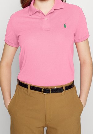 Personne portant un polo rose à manches courtes avec un logo vert brodé et un pantalon beige avec une ceinture noire à boucle dorée.