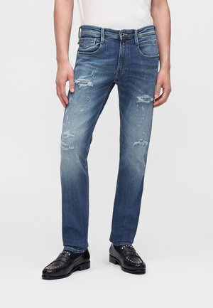 Homme portant un jean bleu usé et des mocassins noirs cloutés, debout les mains détendues le long du corps devant un fond uni.