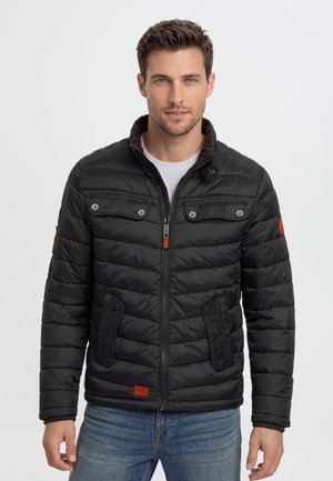 Homme portant une veste matelassée noire avec poches avant et fermeture éclair, associée à un jean bleu, posant devant un fond clair uni.