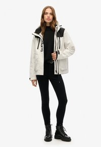 Frau trägt eine schwarz-weiße Kapuzenjacke, einen schwarzen Pullover, schwarze Hose und schwarze Schnürstiefel und steht vor einem einfarbigen Hintergrund.