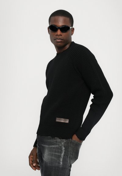 Pull noir en tricot avec un motif texturé, col rond, manches raglan et une étiquette visible. Associé à un jean foncé et des lunettes de soleil noires.