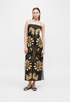 Femme portant une robe noire sans manches avec de grands motifs floraux dorés et des sandales noires ouvertes au niveau des orteils, debout devant un fond uni.