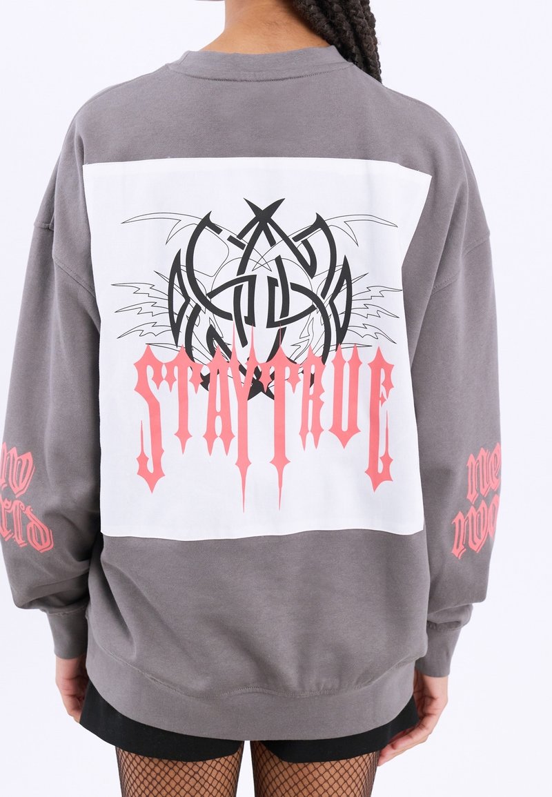 Personne portant un sweat-shirt gris avec un grand patch blanc dans le dos, un design graphique noir, et un texte gothique rose disant "Stature."