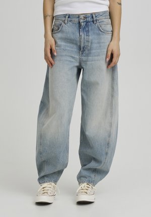 Svetlomodré riflové nohavice s voľným, širokým strihom nohavíc, ktoré majú predné vrecká a zapínanie na zips, v kombinácii s bielymi teniskami.