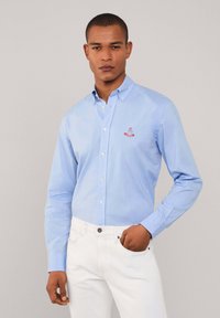 Lichtblauw overhemd van katoen, met lange mouwen, een kraag en een geborduurd logo op de borst. Gepaard met witte broek.