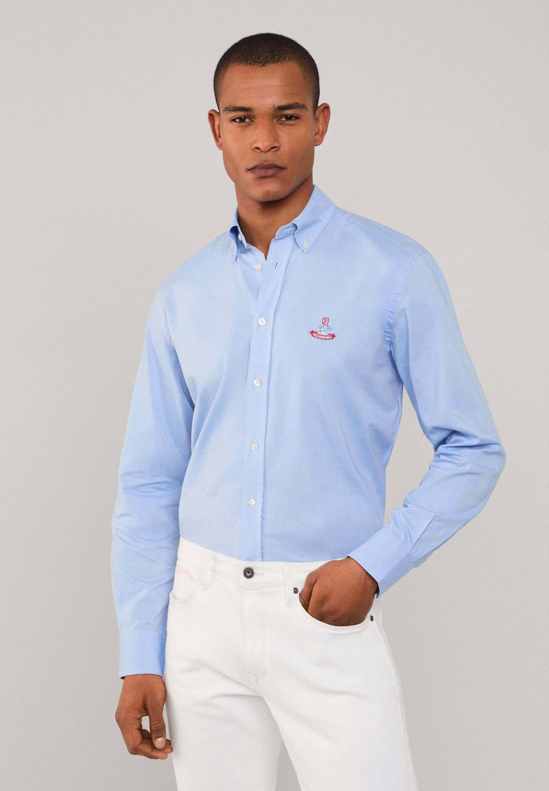 Lichtblauw overhemd van katoen, met lange mouwen, een kraag en een geborduurd logo op de borst. Gepaard met witte broek.