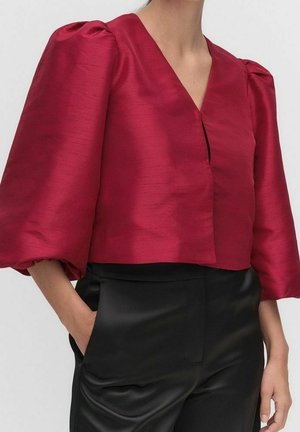 Rode satijnen blouse met driekwart mouwen met pof, V-hals, gecombineerd met hoogwater zwarte satijnen broek, handen gedeeltelijk in de zakken.