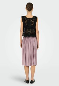 Top in pizzo nero con un motivo floreale, abbinato a una gonna plissettata lilla. Le scarpe sono ballerine nere. Vista da dietro.