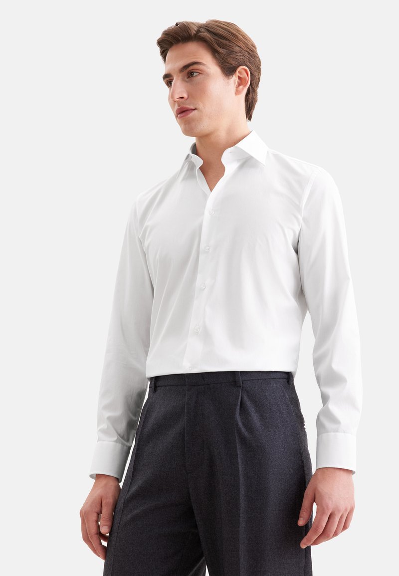 Jeune homme portant une chemise blanche cintrée à manches longues rentrée dans un pantalon gris foncé, regardant légèrement sur le côté sur un fond uni.