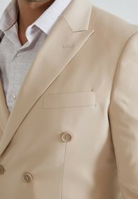Veste de costume beige à double boutonnage avec revers crantés, dotée de deux boutons, d'une poche poitrine et d'un tissu lisse et texturé.