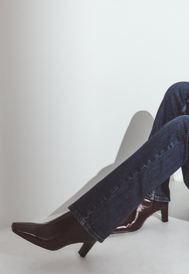 Burgundijas laku ādas augstpapēžu kurpes ar norādītu purngalu un šauru papēdi, kopā ar tumši zilām džinsu biksēm.