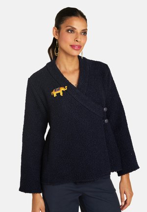 Navy-Wrap-Jacke aus strukturiertem Stoff, mit langen Ärmeln, einem V-Ausschnitt und einem dekorativen goldenen Elefanten-Anstecker auf der linken Seite.