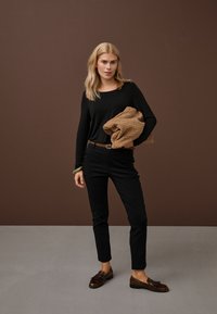 Žena se světlými vlasy má na sobě černý dlouhý rukáv a kalhoty, hnědý pásek a boty, drží složený vzorovaný béžový svetr.