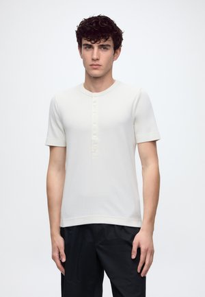 Jeune homme aux cheveux bouclés et foncés, portant un t-shirt henley blanc à manches courtes et un pantalon noir sur un fond blanc uni.