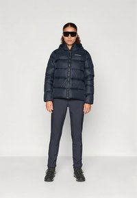 Giacca imbottita blu navy con zip frontale e cappuccio, indossata sopra pantaloni scuri e stivali neri. Modello con occhiali da sole, in piedi dritto contro uno sfondo semplice.
