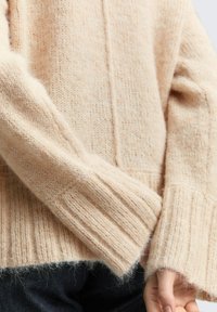 Beżowy sweter na drutach z prążkowanymi mankietami i miękką, puszystą fakturą. Posiada pionowe szwy wzdłuż ciała oraz luźny krój.