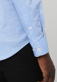 Ljust blå vävd skjorta med långa ärmar, knappmanschetter och kontrasterande vita knappar. Har en standardkrage och böjd fåll.