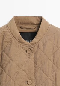 Veste marron matelassée avec col rond et deux boutons visibles, présentant un tissu texturé et un motif de couture en forme de losange.