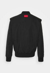 Giacca bomber nera con etichetta rossa al collo, polsini a coste e orlo elasticizzato. Texture liscia e opaca, design aderente.
