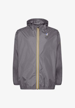 Hombre con chaqueta gris con capucha sobre camisa rosa y camiseta negra, jeans oscuros remangados, zapatos negros sin cordones, sosteniendo un bolso tote negro.