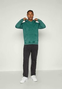Felpa verde in pile con tasca a marsupio e cappuccio con lacci, abbinata a pantaloni neri e sneakers bianche, in piedi contro uno sfondo neutro.