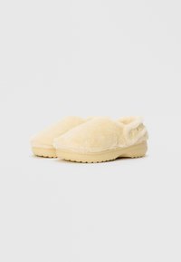 Beige színű, slip-on, bolyhos papucsok, masszív talppal, jobbra dőlt állapotban, állítható, gombos hátsó pánttal.