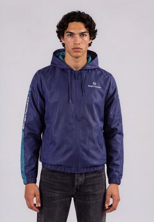 Joven con cabello oscuro y rizado que lleva una chaqueta con capucha azul Sergio Tacchini y vaqueros negros, de pie contra un fondo claro y liso.