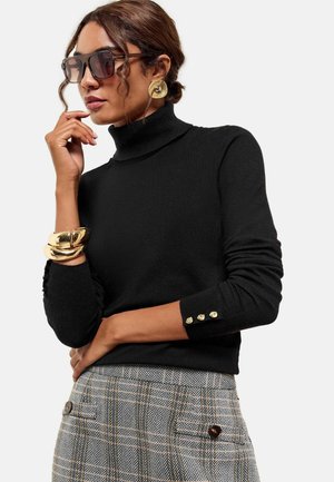 Femme portant un col roulé noir avec des boutons dorés, de grandes boucles d'oreilles dorées, de gros bracelets dorés, des lunettes de soleil surdimensionnées et un pantalon à carreaux taille haute.