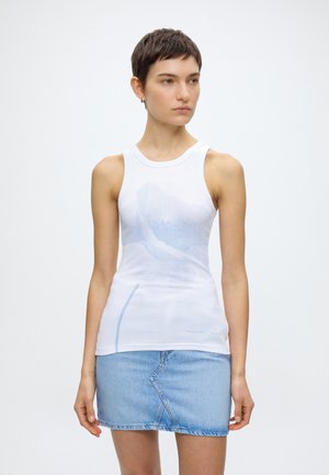 Jeune personne aux cheveux courts et foncés portant un haut blanc sans manches avec un léger motif bleu et une jupe en jean bleu clair, debout sur fond blanc.