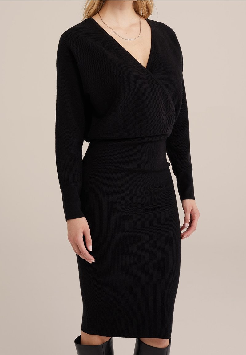 Femme portant une robe en maille côtelée noire à manches longues, un haut cache-cœur à col en V profond, et des bottes noires montant jusqu'aux genoux, sur un fond beige.