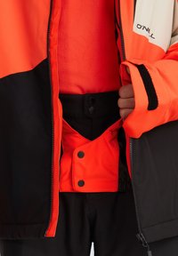 Jasno-pomarańczowa i czarna insulated kurtka z zamkiem błyskawicznym, zatrzaskami oraz widoczną podszewką z polaru. Szczegóły z teksturowanej tkaniny.