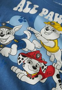 Három animált kutya, kék, sárga és piros egyenruhában, jelvénnyel, mosolyogva és pózolva egy kék háttér előtt, amelyen tappancsnyomok láthatók.