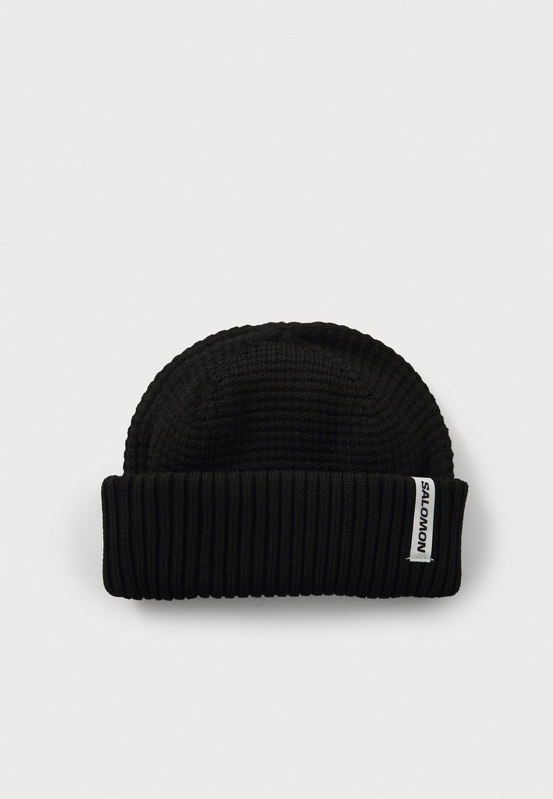 Zwarte gebreide beanie met een geribde manchette en een gladde bovenkant. Bevat een wit etiket met zwarte tekst aan de zijkant. Zachte textuur en flexibele pasvorm.