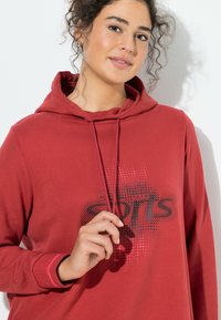 Czerwona bluza z kapturem wykonana z miękkiego materiału, z kapturem na sznurki i czarnym nadrukiem „SPORTS” w kropki na przodzie.