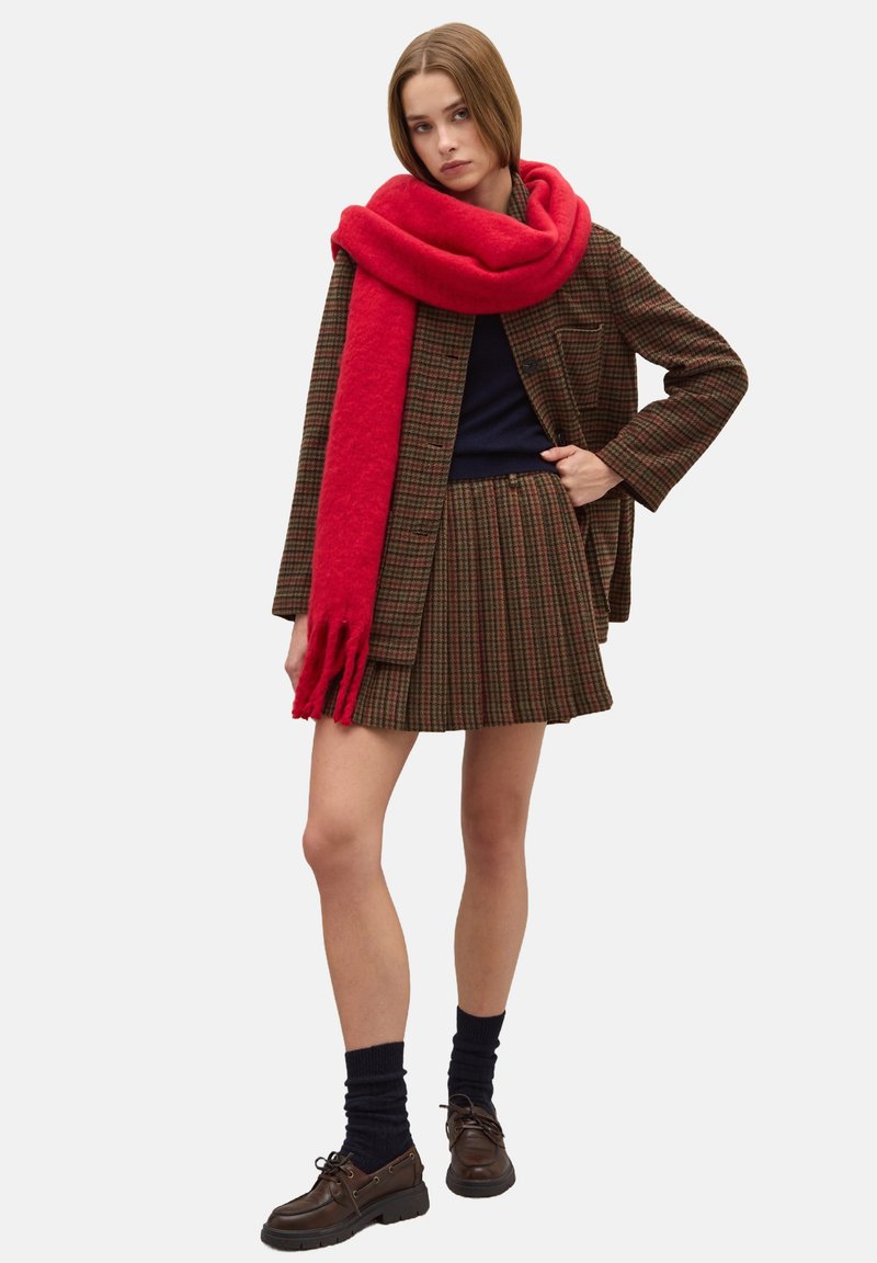 Blazer marrone a quadri sopra una maglia blu navy, abbinato a una gonna plissettata marrone a quadri. Include una sciarpa rossa brillante e scarpe marroni con lacci.