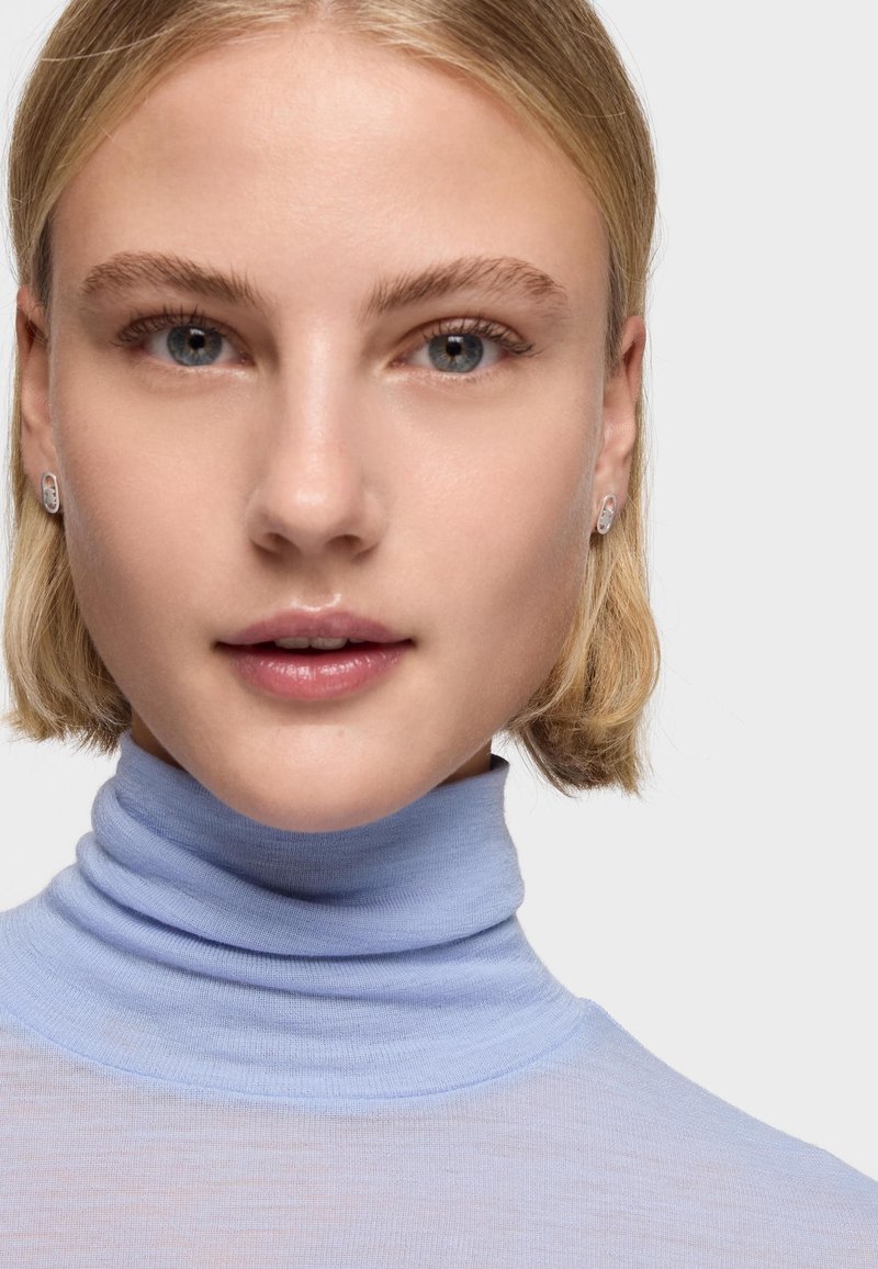 Joven con cabello rubio corto, vestida con un suéter de cuello alto azul claro y pequeños pendientes plateados, mirando directamente al frente con expresión neutral.