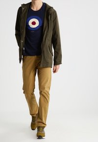 Olivgrön huvajacka, marinblå t-shirt med rund grafik, khakifärgade byxor och bruna sneakers. Texturerna på tyget varierar i hela outfiten.