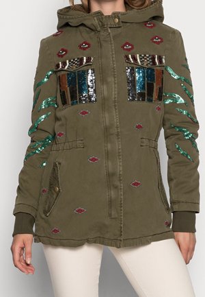 Veste à capuche vert olive avec des patchs en sequins colorés sur la poitrine et les manches, formes losange rouges brodées, portée avec un pantalon blanc.